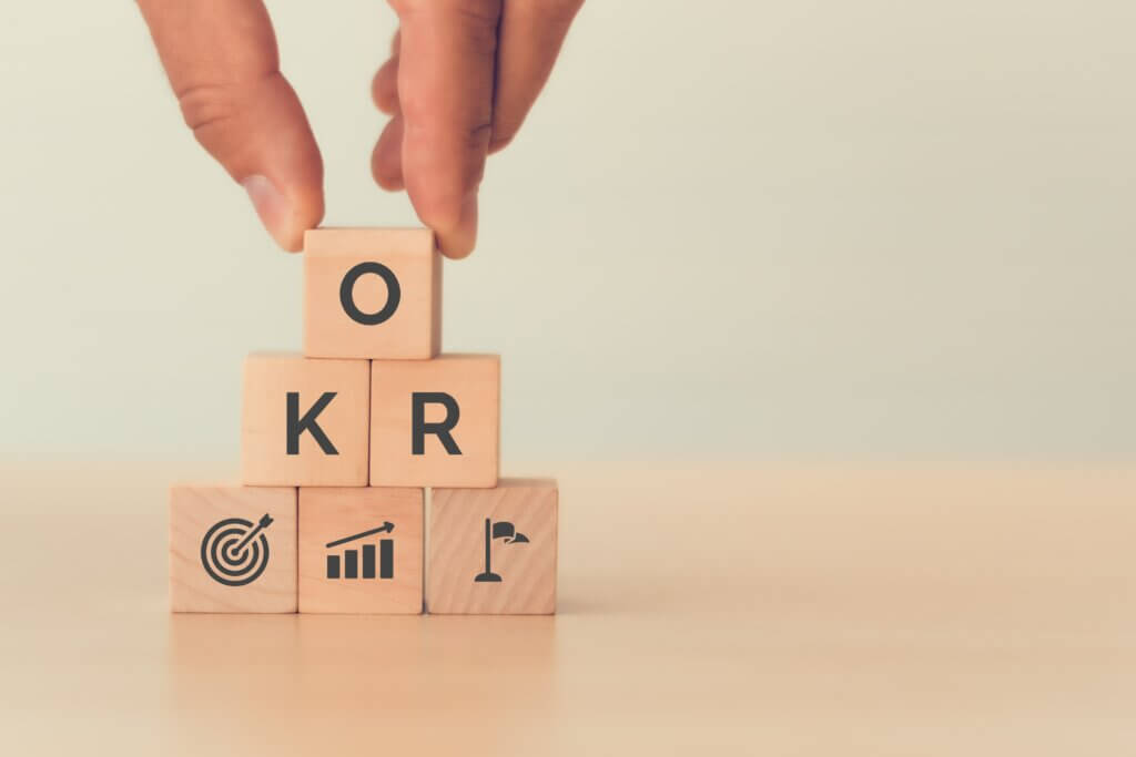 Metodologia OKR, representado por uma mão segurando blocos de cubo de madeira de texto OKR (Objective Key Results) sobre fundo azul. Alvo de negócios e direcionar negócios e desempenho, em busca do alcance do crescimento dos negócios por meio de uma gestão flexível.