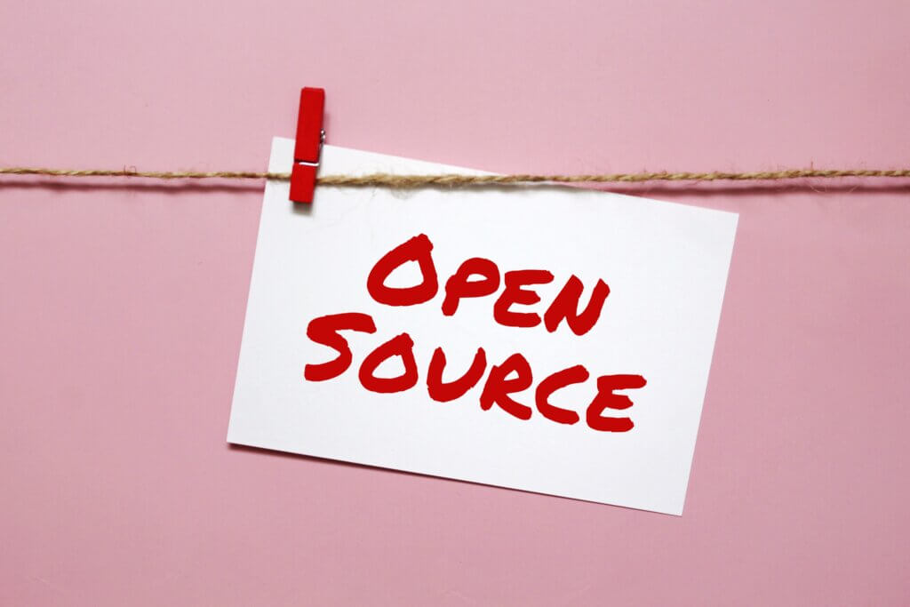 capa artigo projeto open source. Uma foto com fundo rosa e um papel em branco escrito 'open Source" em vermelho preso em uma corda com um pregador vermelho.