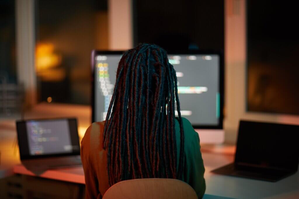 Capa do artigo sobre Java 17 onde vemos uma pessoa negra com dreadslocks compridos de costas, a sua frente há duas telas, um monitor e um notebook onde é possível identificar códigos de programação.