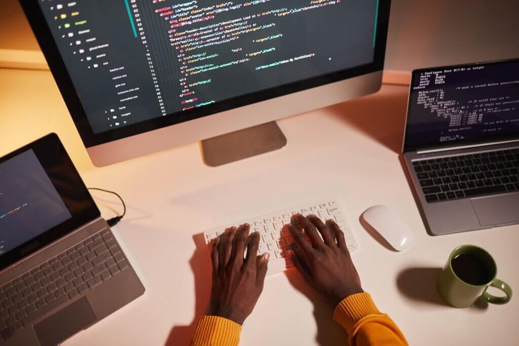 Capa do artigo Redux + Redux Saga: gerenciamento de Requisições React onde vemos mãos digitando em um teclado sem fio com um monitor a sua frente, dois notebooks e um mouse ao lado