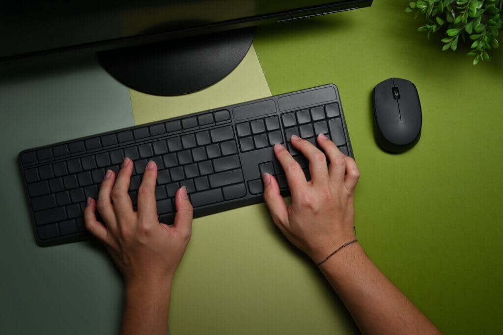Capa do artigo sobre Documentação de Software em que vemos mãos de uma pessoa jovem digitando em teclado sem fio preto e um mouse preto, também sem fio. A mesa é pintada com três tons diferentes de verde e ainda podemos ver uma planta no canto superior direito.