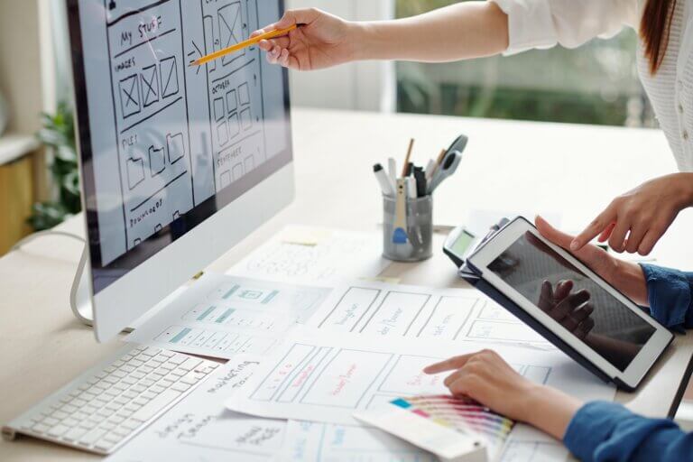 Capa do artigo sobre Product Sense, onde duas pessoas estão a frente de um computador, tablet e alguns rascunhos em papel na mesa de trabalho.