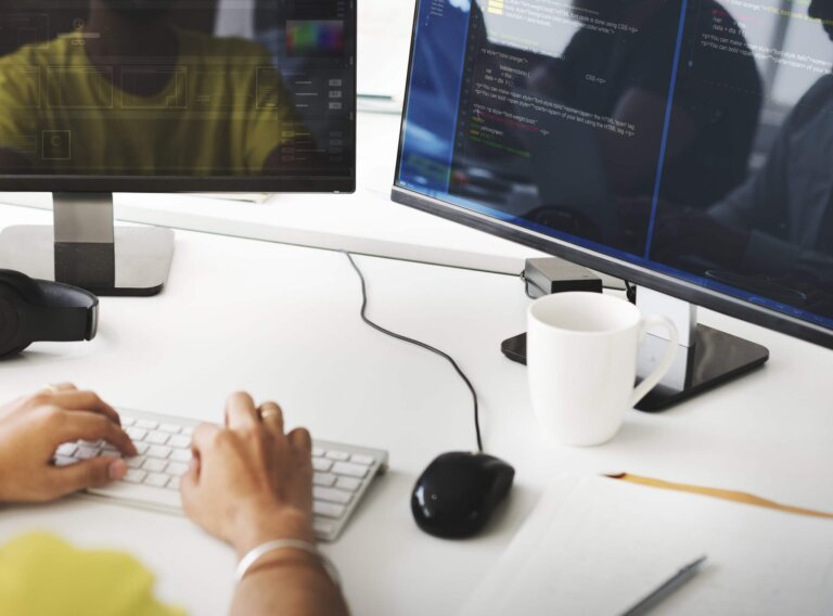 Capa do artigo sobre o que é um bom código para Programas de Formação de pessoas desenvolvedoras, onde temos uma pessoa trabalhando com código em duas telas, junto com um teclado e mouse.