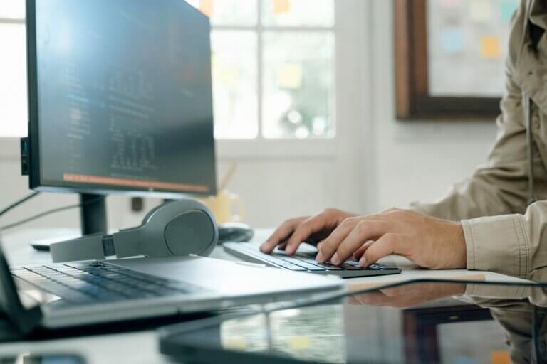 Capa do artigo sobre boas práticas de GitHub Actions. Na foto, uma pessoa está digitando em seu teclado, aparecendo somente parte do corpo. O resto da imagem é composto por um monitor, um headphone e uma mesa, ao fundo está uma janela.
