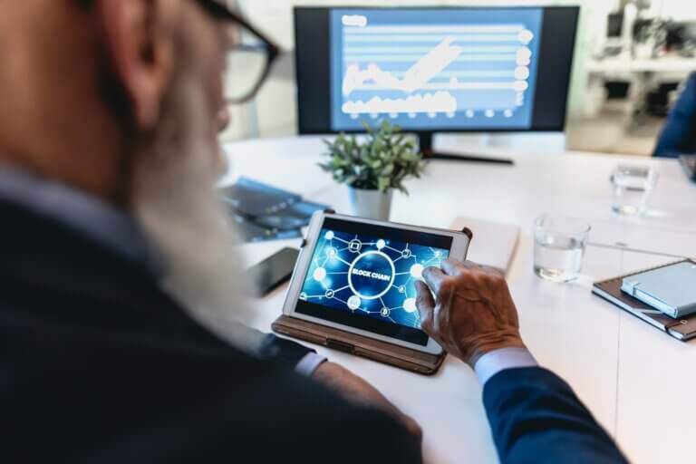 Capa do artigo sobre DeFi ou finanças descentralizadas. Na imagem, um homem branco de barba branca e óculos segura um tablet com a palavra blockchain.