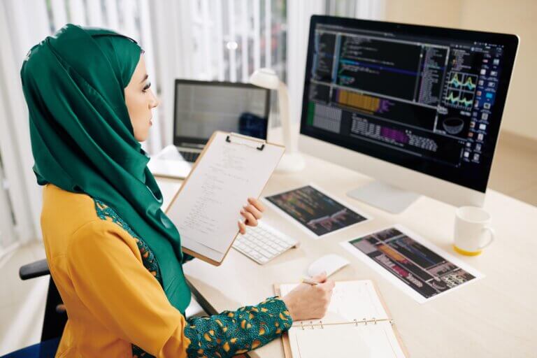 Capa do artigo sobre Stack Overflow Survey 2022. Mulher com hijab está em frente ao computador com códigos segurando uma prancheta.