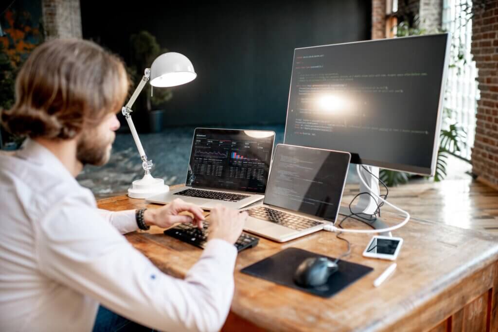 Capa do artigo sobre docker na prática. Na imagem, um jovem programador do sexo masculino escrevendo um código de programa sentado no local de trabalho com três monitores no escritório.
