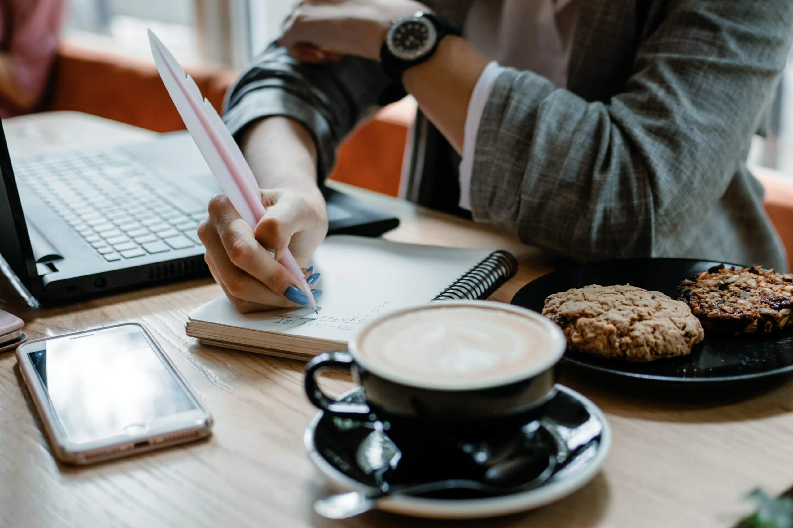 Mulher branca sentada em uma mesa em frente a um notebook, com uma xícara de café, um cookie e seu celular, dá início ao seu questionário de pesquisa em um caderno de anotações