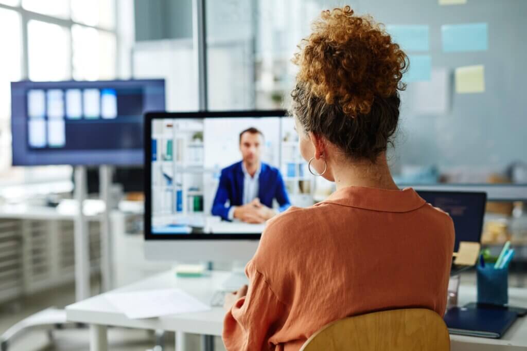 Mulher sentada de frente para uma tela de computador aplicando uma reunião one-on-one ao seu liderado. Na tela do computador está um homem, com paletó e camisa de botão.