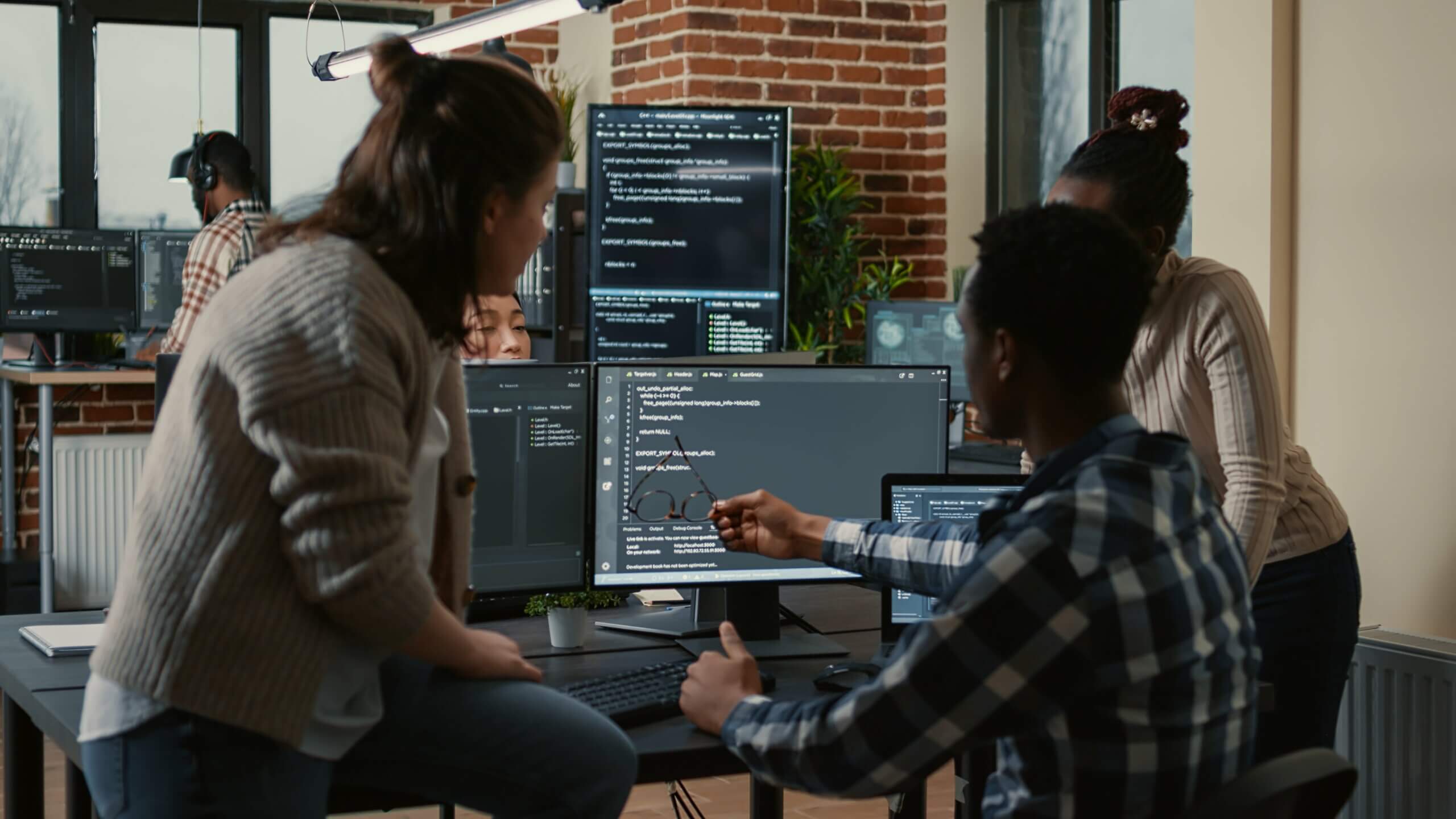 Essa imagem ilustra o conteúdo "Gerente em desenvolvimento de software: quais são as melhores características", onde pessoas programadoras estão sentadas na mesa discutindo com equipe mista de pessoas desenvolvedoras de software. Na imagem é possível ver 4 telas com código.