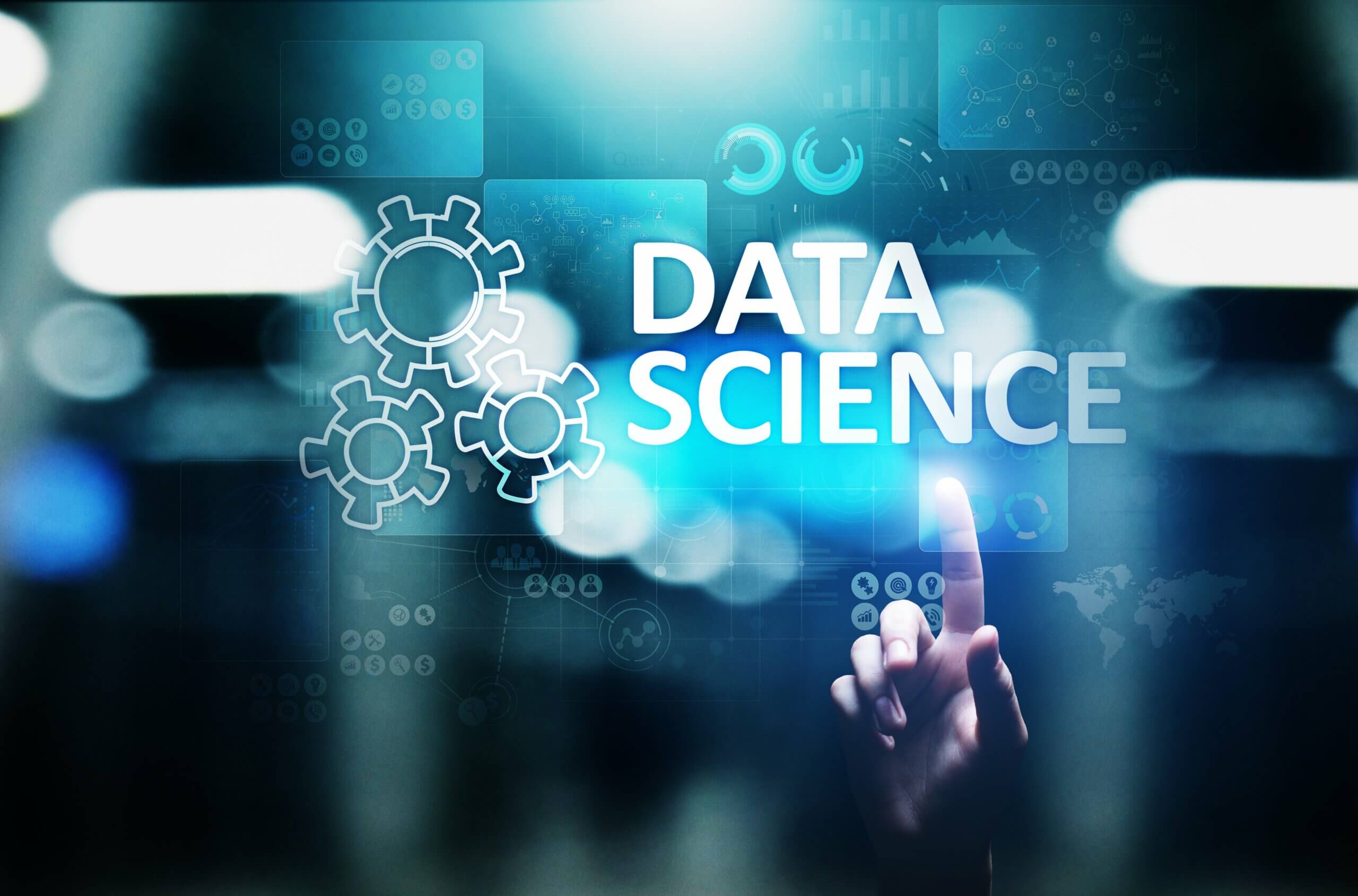 Imagem com “Data Science” em destaque em um fundo azul com elementos que remetem a dados, como engrenagens e gráficos. Apontando para o título, está uma mão de uma pessoa branca.
