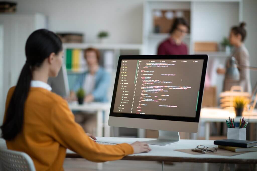 Imagem capa do conteúdo ECS Fargate, onde uma mulher branca está de frente para o seu computador na sua empresa. A tela do dispositivo mostra códigos e ao fundo, colegas de trabalho.