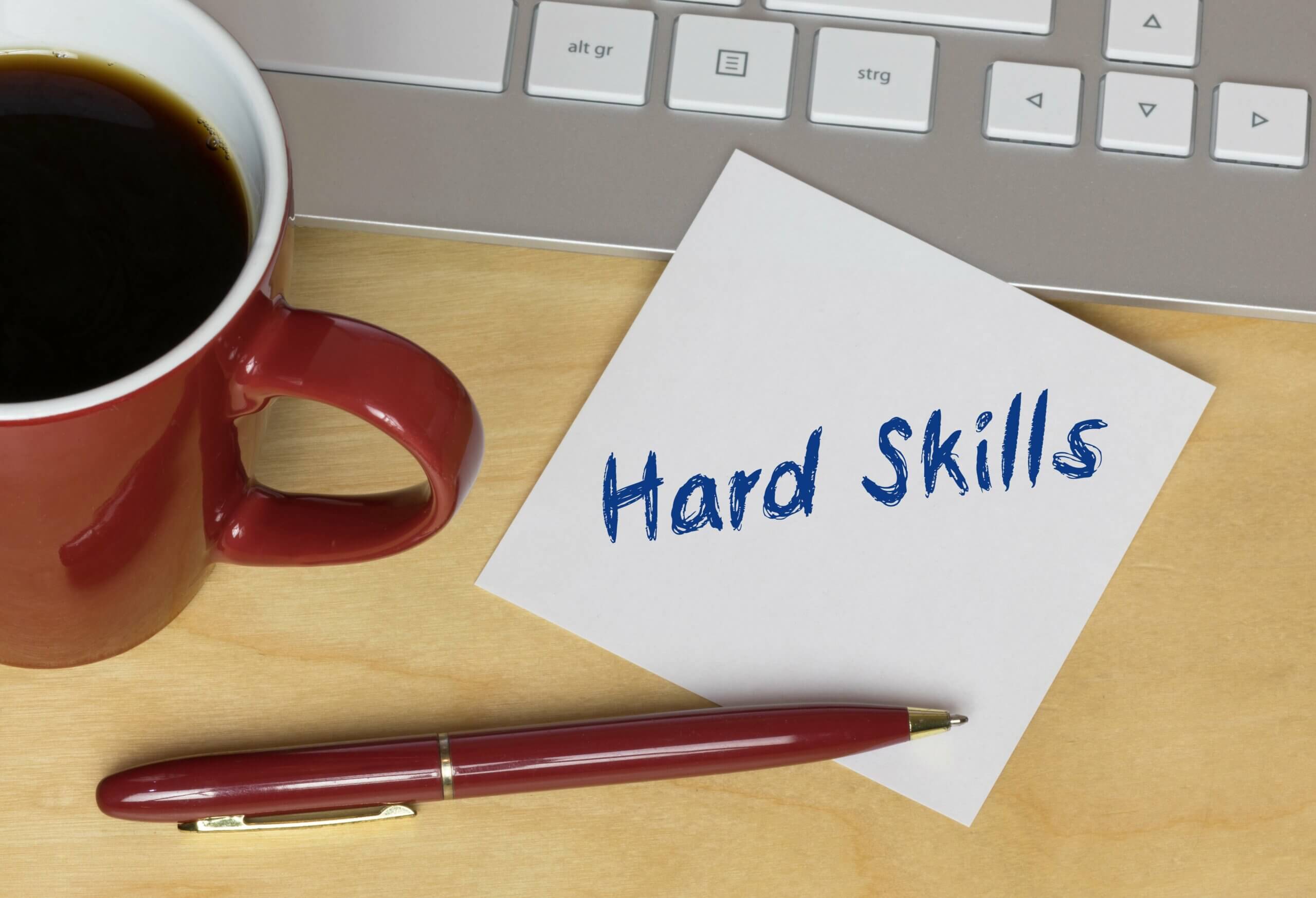 Imagem capa do conteúdo sobre "Mapeamento da Hard Skills", onde em uma mesa de madeira podemos ver uma caneca cheia de café, um teclado de computador, uma caneta com uma metade apoiada na mesa e a outra em um papel, escrito "Hard Skills"