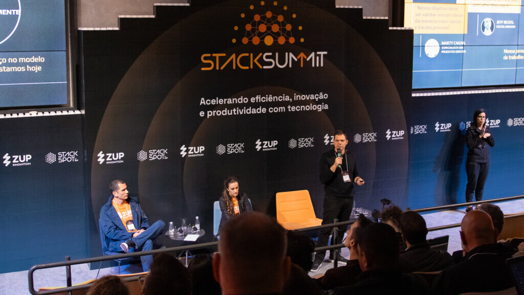 Foto de três pessoas durante a agenda StackSpot e Generative AI, André Palma, Manuela Leoni e Fabio Napoli. Todos estão no palco, Fabio está de pé e os outros sentados, em frente ao painel StackSummit.