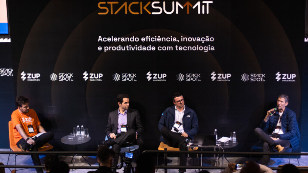 Foto de quatro homens durante a agenda Salesforce com Caio Jardim, Gabriel Dornella, André Piotto e Jaime Müller. Todos estão no palco, sentados, em frente ao painel StackSummit.