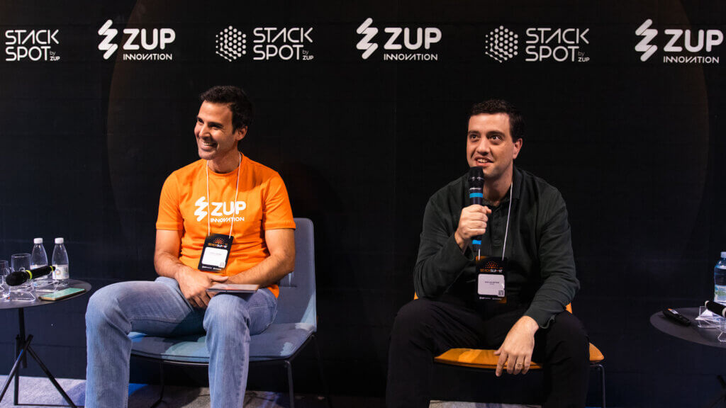 Foto de dois homens na agenda iFood, Alberto Souza e Igor Hjelmstrom, no palco do StackSummit, ambos estão sentados e atrás está o painel do evento.