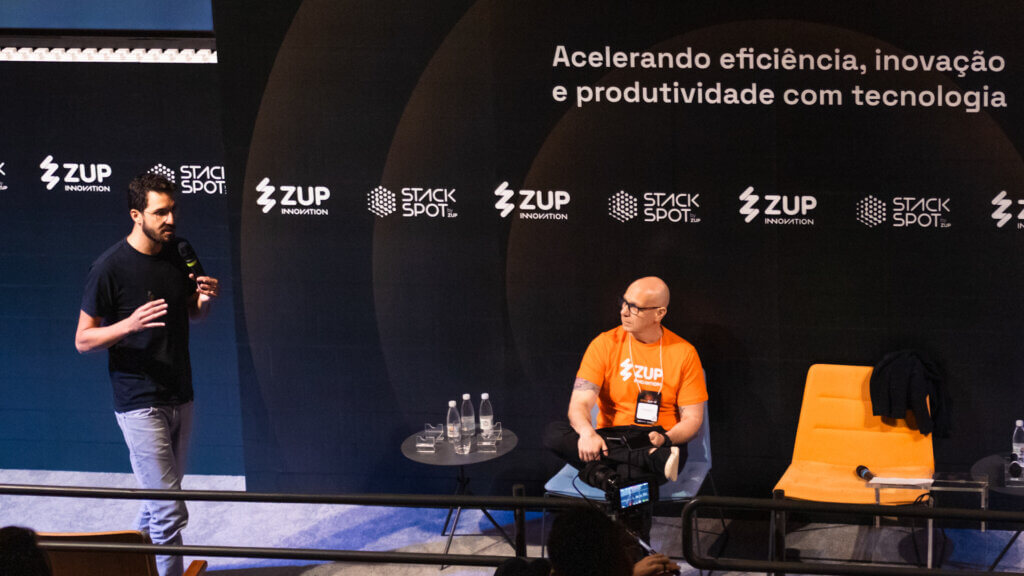 Foto de dois homens na agenda sobre IA, Renato Barbosa e Carlos Ferraiuolo, no palco do evento. Atrás deles está o painel do StackSummit em preto com o tema e os promotores: Zup e StackSpot.